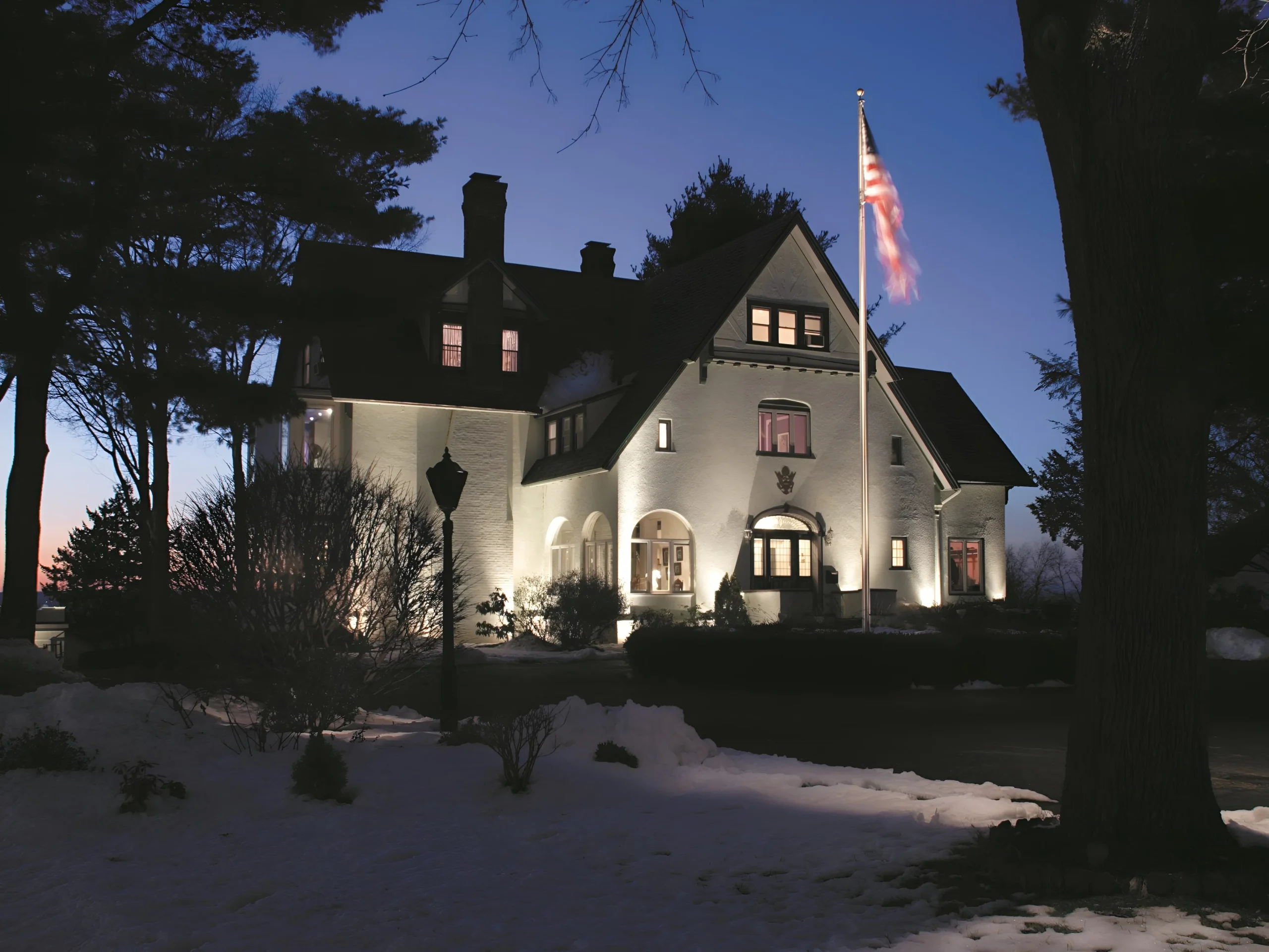 Uplights wash a white stucco house above snow, lighting maintenance for Scarsdale homes.