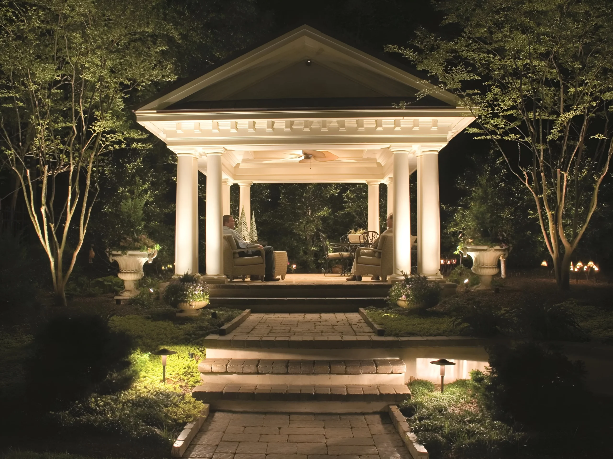 Two people sit in a lit, columned garden pavilion framed by uplit trees at night.